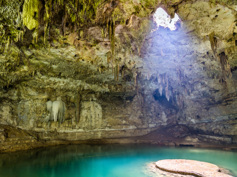A cenote with light streaming through, showing beautiful turquoise water and many stalactites.